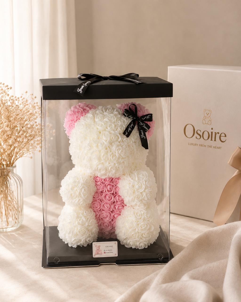 Figura de oso cubierta de rosas preservadas claras, bajo cúpula, junto a su caja de empaque.